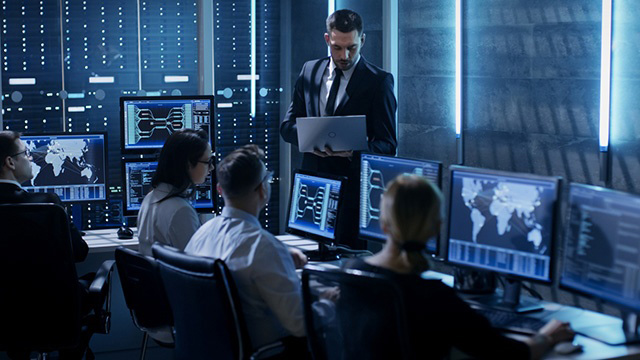 Professional IT Engineers Working in System Control Center Full of Monitors and Servers. Supervisor Holds Laptop and Holds a Briefing. Possibly Government Agency Conducts Investigation.